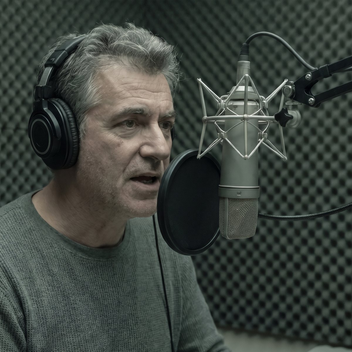 A gray-haired man in headphones speaks into a studio microphone with a pop filter, in a booth lined with foam panels.